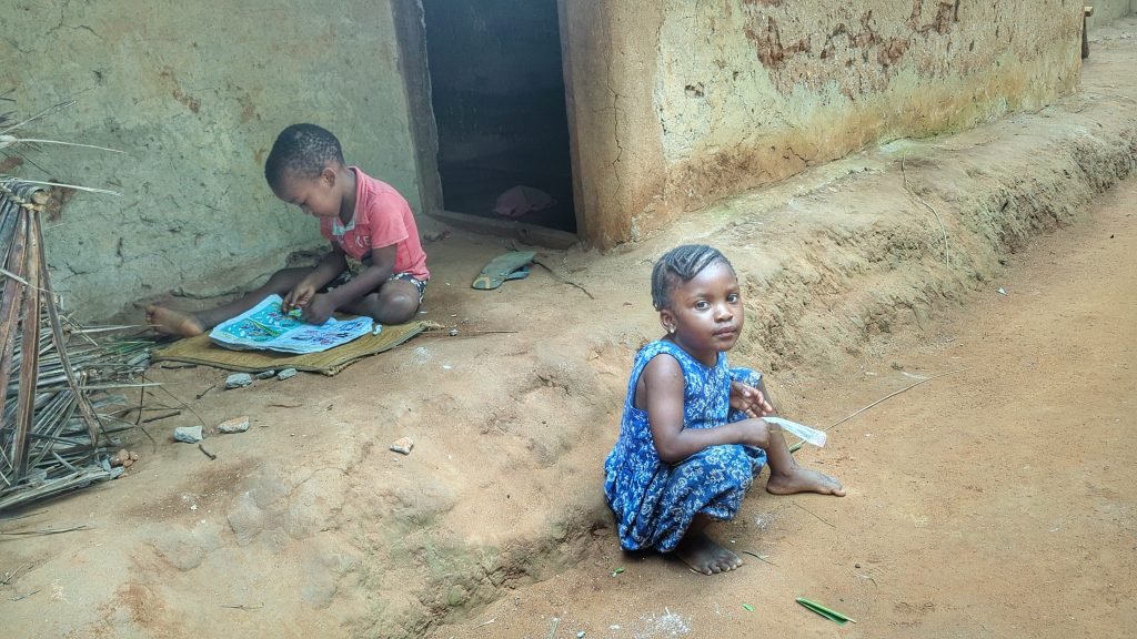 Zwei afrikanische Kinder sitzen vor einem einfachen Haus, eines liest, das andere schaut nachdenklic.