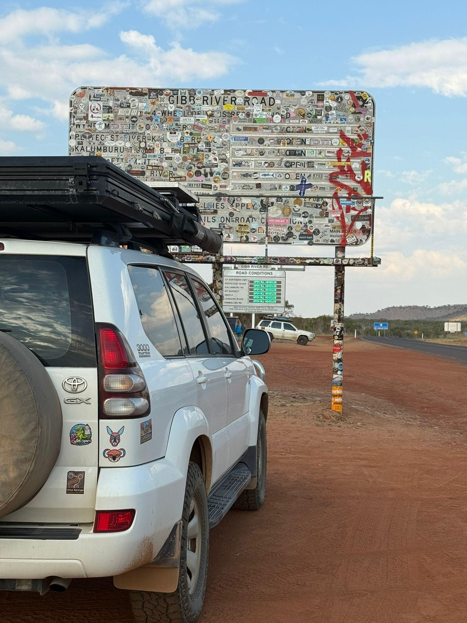 Geländewagen mit Dachbox vor einem großen, bemalten Straßenschild in Australien.
