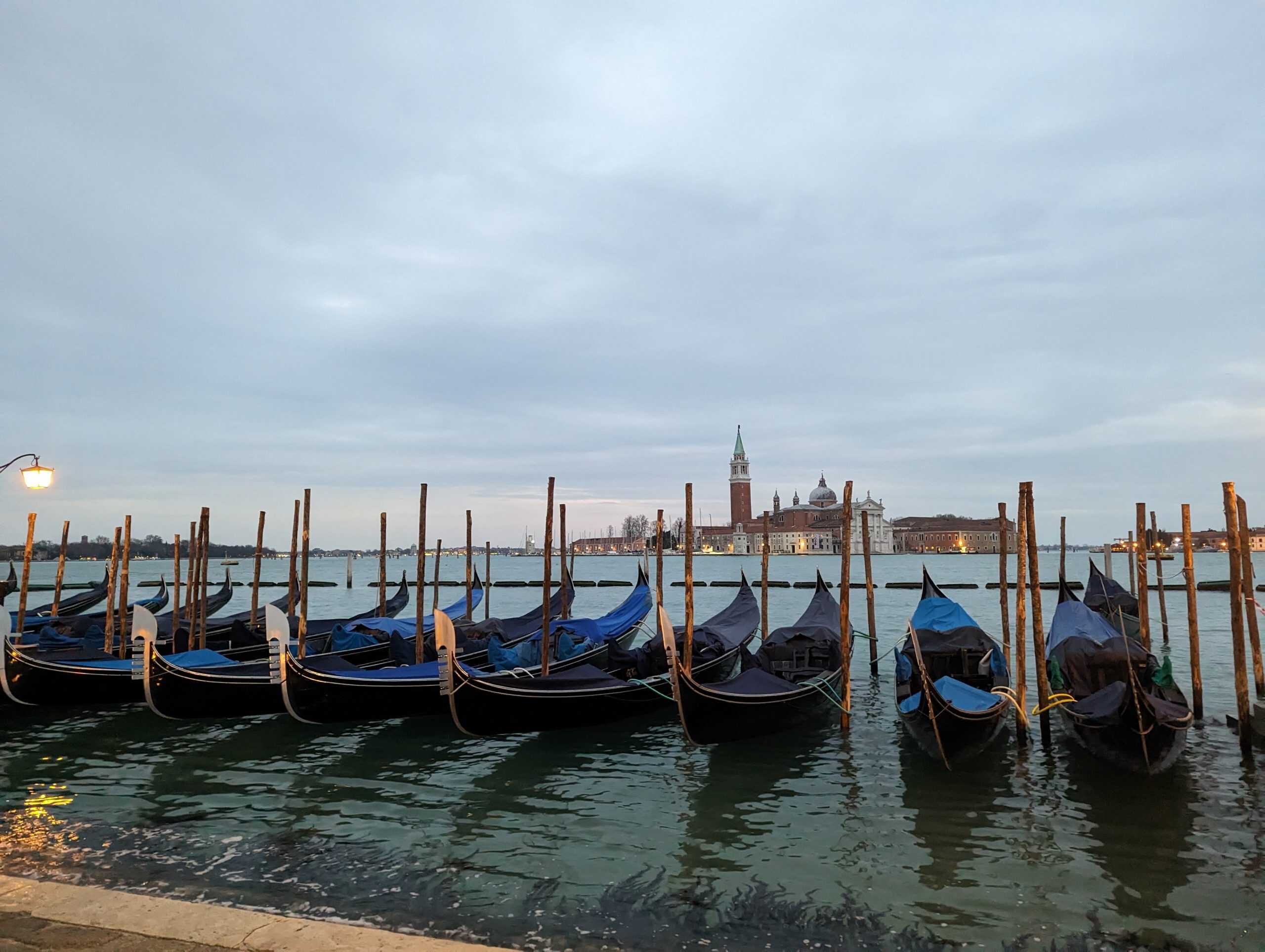 Gondeln in Venedig, ruhiges Wasser, historische Stadt im Hintergrund, bewölkter Himmel.